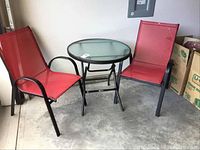 Two red mesh chairs with black metal frames flanking round glass top table with black metal legs on concrete floor.