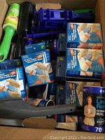 Photo of a box filled with various Lintek lint rollers in blue and white packaging along with some black and green lint roller handles and blue plastic holders.