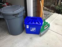 Rubbermaid Brute grey garbage can, Home Depot blue recycle bin, and Garent leaf rake side by side on concrete floor near garage.
