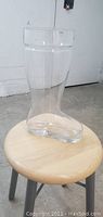 Close-up view of one clear glass beer boot with rounded toe on a wooden stool against a white background.