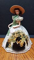 Front view of the Gone With The Wind porcelain figurine showing detailed painting on the skirt with movie scenes and the woman in the picnic dress wearing a hat and green scarf.