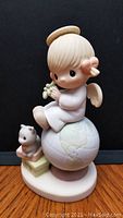 Front view of Precious Moments angel porcelain figurine sitting on a globe holding a flower, with a teddy bear beside it.