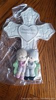 Photo of the figurine wrapped in plastic, showing a white cross with embossed arrow details and a heart inscribed with 'Happy 50th Anniversary' with a boy and girl figurine kneeling.