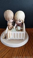 Front view of porcelain bisque Precious Moments figurine depicting two children with a baby in crib and teddy bear, on round base.