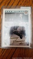 Front view of autographed Johnny Bower hockey card in protective sleeve on wooden surface.