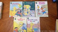 Five Sesame Street hardcover books arranged in two rows on a wooden floor; top row has three books and bottom row has two. Each book has a colorful cover featuring Sesame Street characters.
