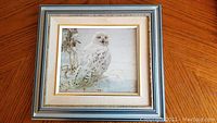 Framed picture of a snowy owl perched on a branch with natural background, silver and gold-tone frame with cream-colored matting.