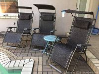 Three zero gravity lounge chairs with black metal frames and patterned fabric, each with headrest pillow and sunshade canopy, plus a small light blue metal side table on an outdoor patio.