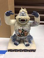 Front view of Bumble the Abominable Snow Monster figurine with painted images of Rudolph and Hermey on the front belly, standing on snowy base.