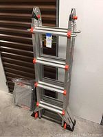 Collapsed ladder and accessories leaning against storage door, platform and paint tray in bag on floor