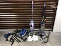 Three cleaning appliances including a purple stick vacuum, a blue canister steamer with accessories, and a Shark Duo Clean upright vacuum with attachments laid out on floor.