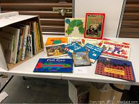 Photo of the table with all books laid out showing covers and titles with a box of additional books in the background.