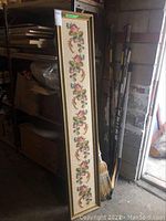 Full view of framed long rectangular needlepoint artwork leaning against boxes and broom in a storage area.