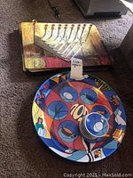 Photo showing silver-tone Menorah with nine candle holders on top of placemats, next to colorful round Passover Seder plate with matching small bowls.