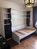Full view of assembled white and black single bed frame with trundle and attached shelving and drawer units in room.