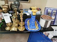 Shelf with six brass candlesticks arranged alongside a blue painted tin tray holding two blue and yellow candle holders and a round decorative tin container.