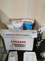 Three plastic yogurt tubs in Luxardo cardboard box on table with O Canada sheet underneath