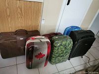 Photo showing front view of 6 luggage pieces lined up including the red maple leaf hard shell, green camouflage hard shell, brown, maroon, black and blue soft shell suitcases.