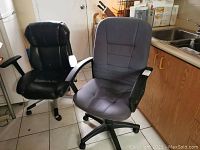 Two office chairs side by side: A black faux leather chair and a gray fabric chair with plastic armrests in a kitchen setting.