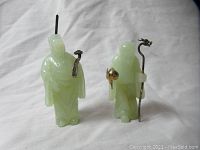 Both figurines shown together on white cloth background, highlighting their jade material and metal accessories.