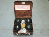 Open brown carrying case holding four black Henselite bocce balls with green rings and gold lettering, white cloth, and accessories.