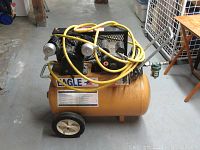 Front view of compressor showing orange tank, two wheels, black pump assembly and yellow hose draped over top.