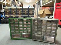 Three plastic drawer organizers of blue, green, and gray colors arranged on floor in a workshop setting, containing small hardware parts.