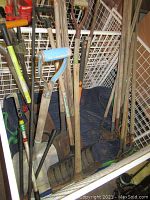 Overhead view of assorted garden tools including shovels, hoes, and rakes with wooden handles against a wire fence background