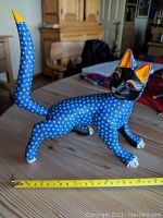 Front and left side view of blue Alebrije wooden cat sculpture showing detailed dotted painting and raised tail with yellow tip, sitting on wooden table with measuring tape for size reference.
