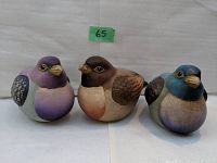 Front view of three ceramic birds side by side, showcasing hand-painted details and colors: purple, brown, and blue.