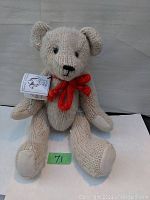Front-facing image showing cream-colored knitted teddy bear with black embroidered facial features and red knitted bow, seated with arms and legs articulated.