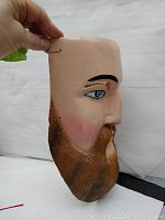Side profile of hand carved wooden mask showing detail of carved beard, blue eye, and skin tone paint.