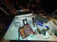 Overview of all items including the scale, knife, spoon, purses, lamp, and fly box on white table.