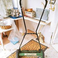 Green Lee Valley manual push reel lawn mower standing indoors on a doormat with a visible 18-inch blade reel and black handle grips, lightly dusted but in good condition.