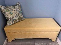 Front and side view of the ultra sued fabric storage bench box with nailhead trim placed in a corner with a cushion on top.