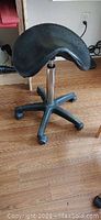 Black saddle-shaped stool with leather-like seat, adjustable height, and wheeled base on wood floor.