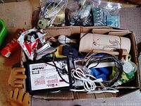 Top view of a cardboard box containing various electrical components including a Shur Shock fence controller, cables, outlet extenders, lamp parts, and a wooden tool.