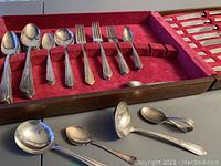 Full view of the flatware in the storage box showing spoons, forks, and ladles in red fabric-lined wooden box.