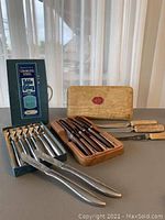 Three sets of Sheffield knives displayed: one blue box with stainless steel knives, one beige box with wooden-handled knives, and three extra wooden-handled knives showing wear