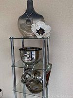 Chrome decorative shelf unit showing two chrome bowls, a chrome plate, large metallic vase, and white floral accent