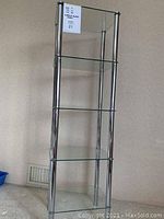 Full view of the tall shelf unit showing the four glass shelves and chrome frame, against a plain wall.