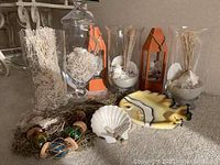 Several clear glass vases with natural beach elements, two orange lantern candle holders, decorative shells, a fish-shaped plate, and sea glass floats arranged on a surface