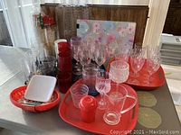 Overall view of used plastic serving ware including clear and colored plastic wine glasses, red trays, clear pitcher, small plastic containers, and floral-patterned trays in the background.