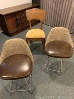 Shows two matching mid-century style bar stools with metal legs, brown faux leather seats, and woven fabric backs, along with wooden chair and side table in background