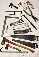 Photo showing various used hand saws, two axes, caulking guns, pruning shears, metal square tool, clamp, and crowbar laid out on a flat surface.