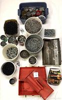 Photo showing multiple white buckets, small containers, and bins with assorted nails and screws in various sizes, along with metal tool trays and a red plastic organizer.