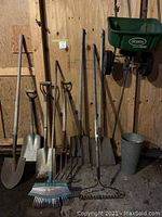 Photo of various gardening tools lined up against plywood wall including shovels, rakes, pitchfork, dolly, lawn spreader and bucket.