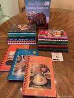 Full view of all 14 cookbooks stacked in two piles on a table along with some spiral-bound Company’s Coming cookbooks laid flat in front. Includes colorful covers featuring food photography.