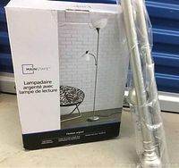 Boxed Mainstays silver floor lamp with reading light shown next to wrapped silver curtain rod.