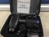 Black massage gun in its hard carrying case with user manual and multiple interchangeable massage heads, power cable connected and lit LED power indicator visible.